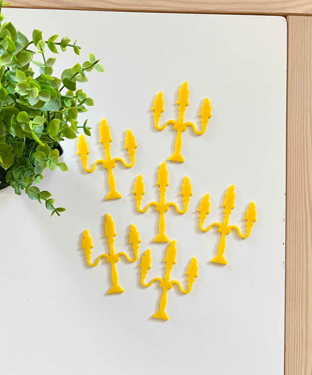 Six bright yellow candelabra-shaped cutouts arranged on a white surface next to a green leafy plant