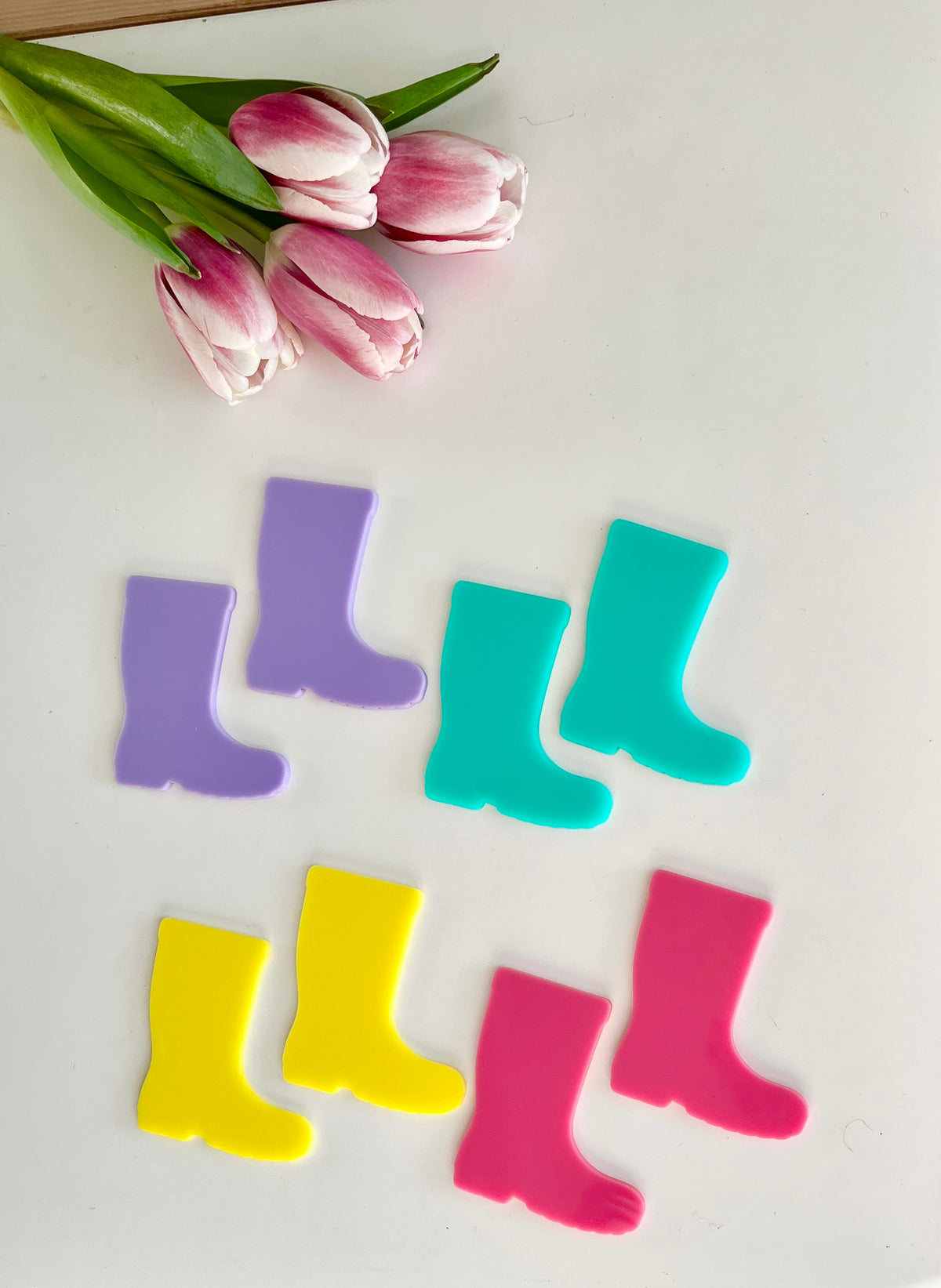 Four pairs of colorful rain boot cutouts in purple, blue, yellow, and pink beside pink and white tulips on white surface
