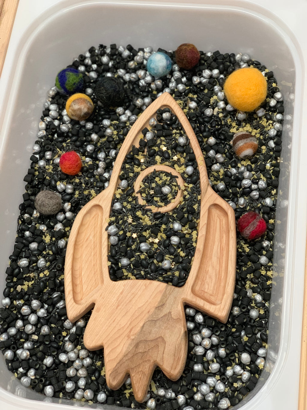 Wooden rocket-shaped tray surrounded by black, silver, and gold sensory beads with colorful felt balls resembling planets