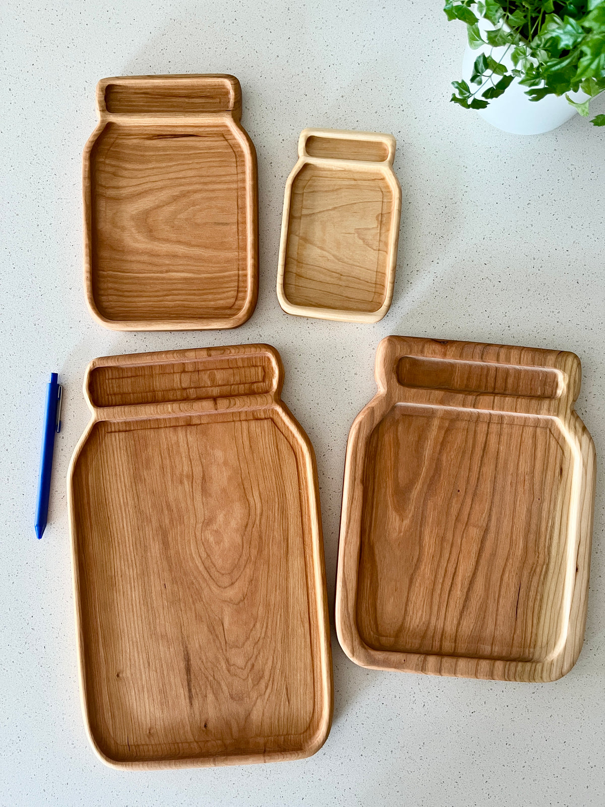 Set of four wooden trays shaped like jars in varying sizes placed on a white countertop near a green plant and blue pen