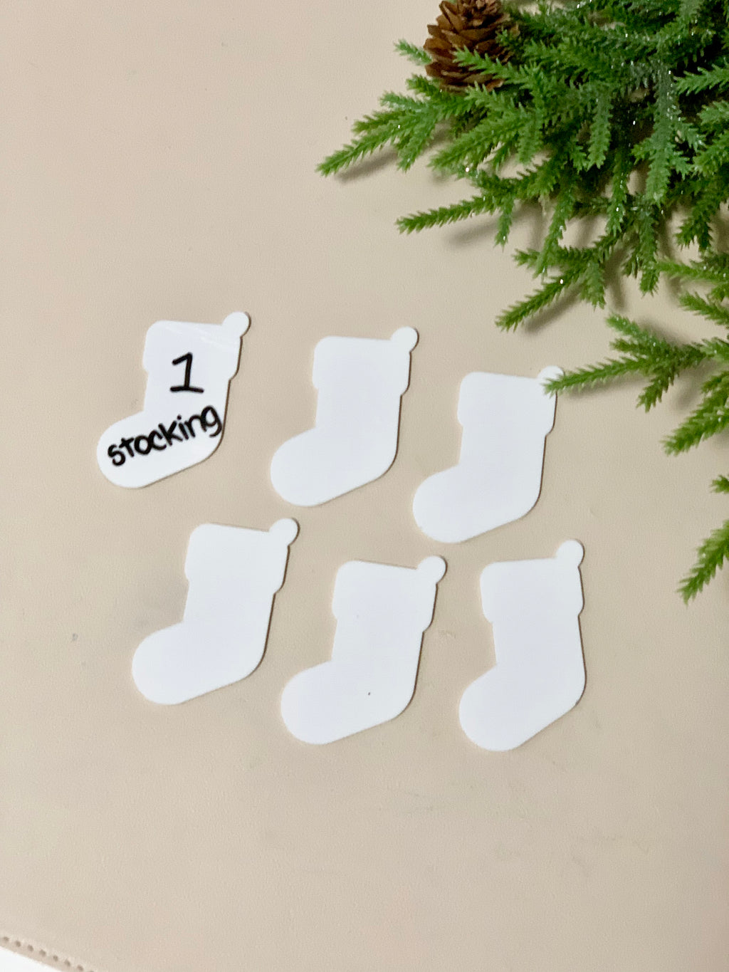 Set of six white dry erase Christmas stocking shapes on beige surface with green pine branch in corner