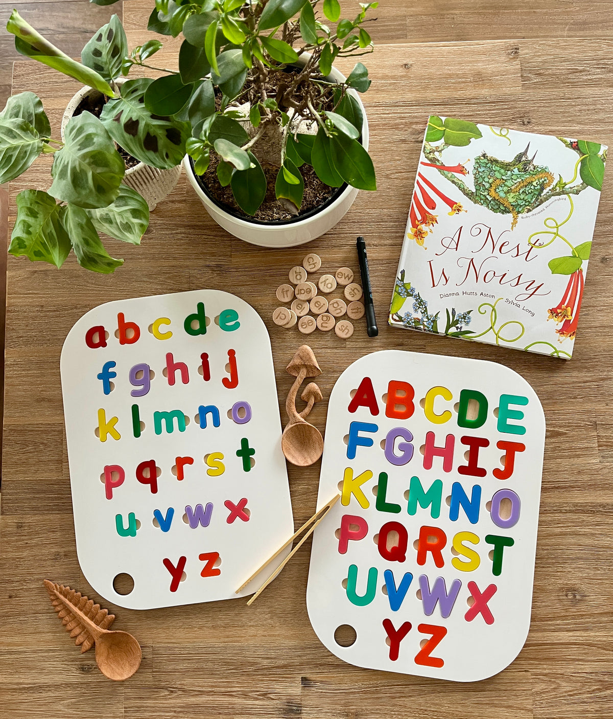 Two Acrylic Dry Erase Lowercase Alphabet Puzzle Flisat Table Top Inserts with Both Rainbow and White Letters arranged on a wooden surface with wooden scoops, alphabet discs, a pen, plants, and a nature-themed book for handcrafted homeschool education resources.