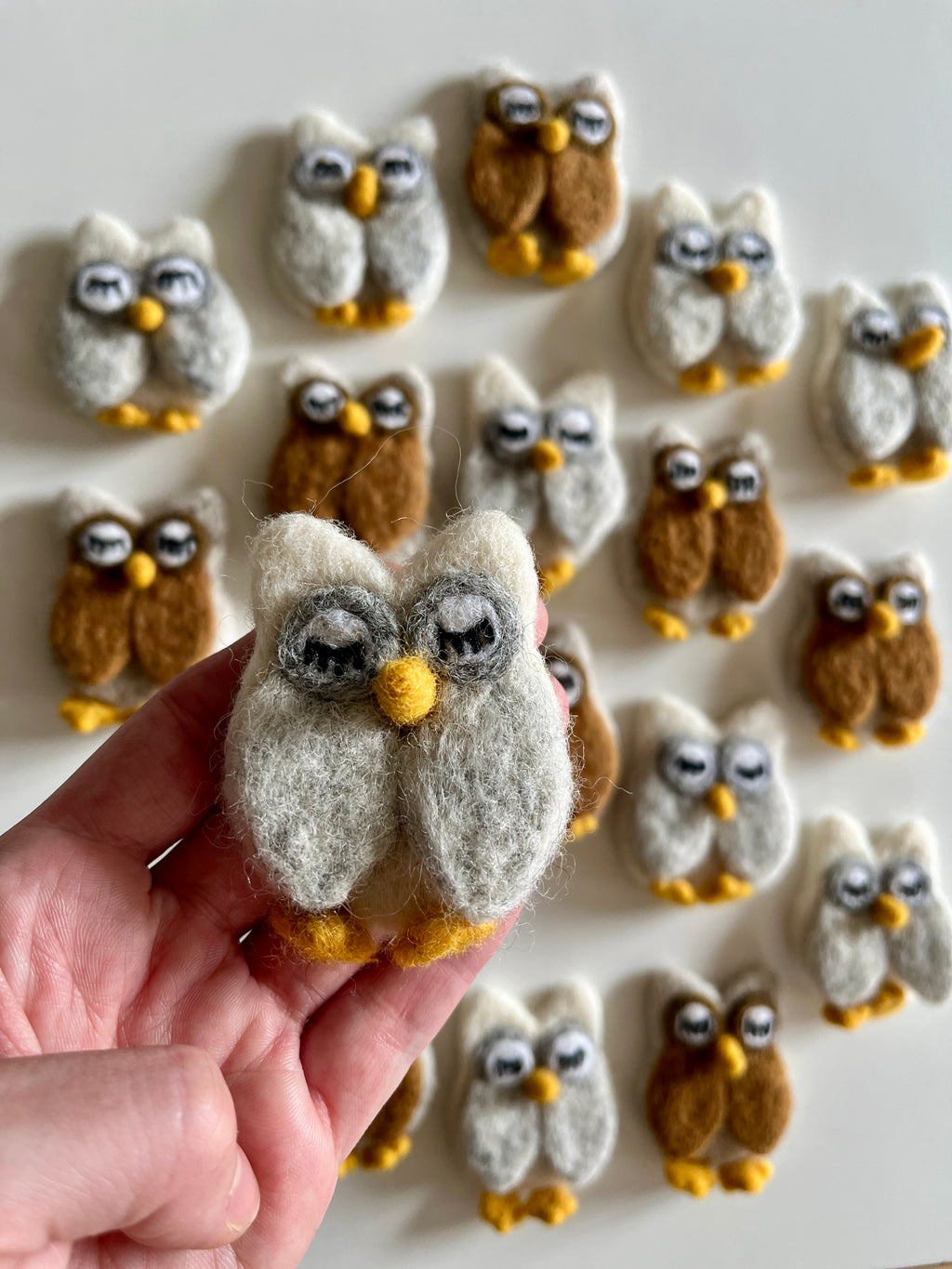 Hand holding a small felt owl figure in gray and white with more similar brown and white felt owls arranged on white surface behind