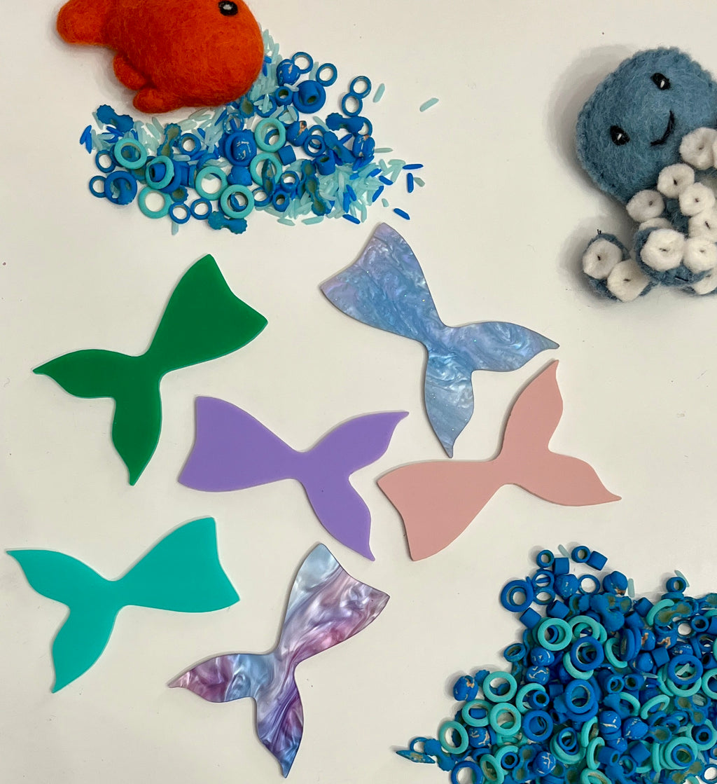 Assorted colorful acrylic mermaid tails with blue and teal ring-shaped pieces and felt ocean creatures on white surface