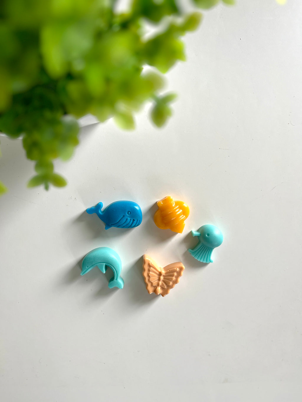 Five small colorful animal and insect-shaped figures arranged on a white surface next to green plant leaves