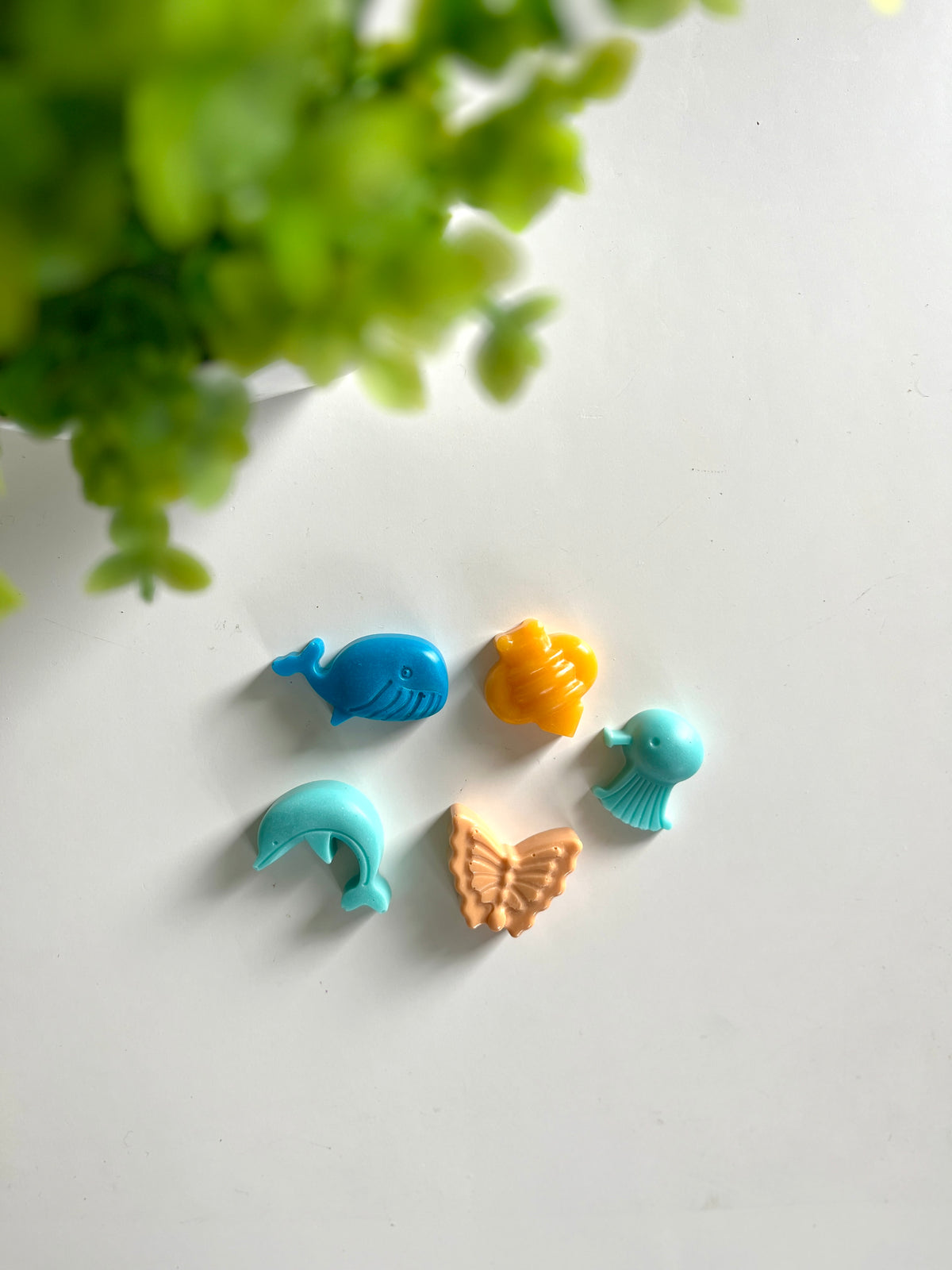 Five small colorful animal and insect-shaped figures arranged on a white surface next to green plant leaves