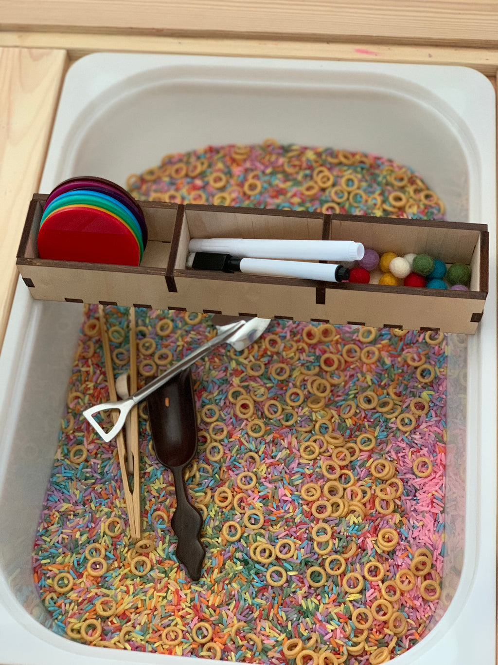 Sensory bin filled with colorful rice and cereal rings, featuring wooden tools, small felt balls, markers, and colored discs in a tray organizer