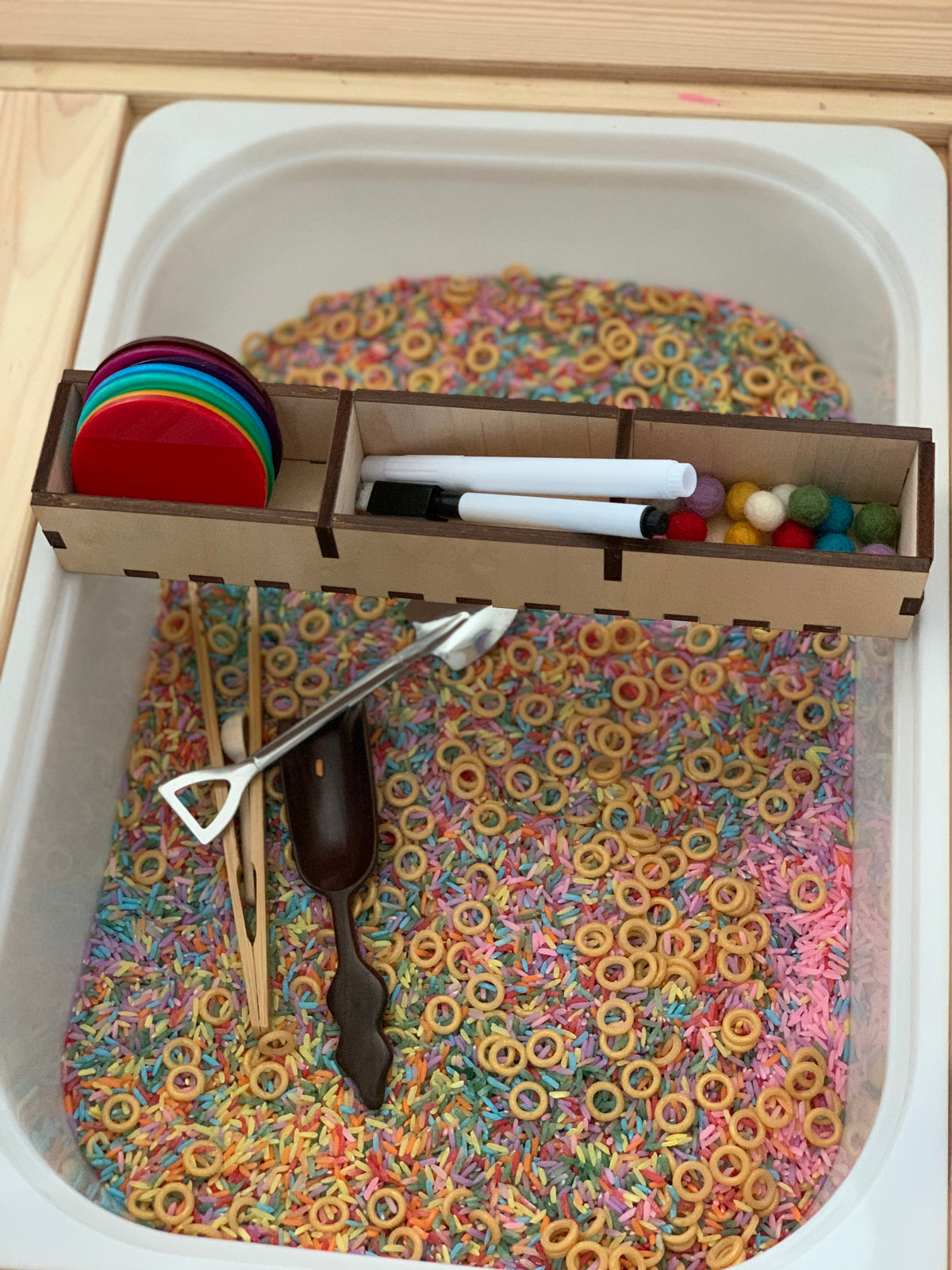 Sensory bin filled with colorful rice and cereal rings, featuring wooden tools, small felt balls, markers, and colored discs in a tray organizer