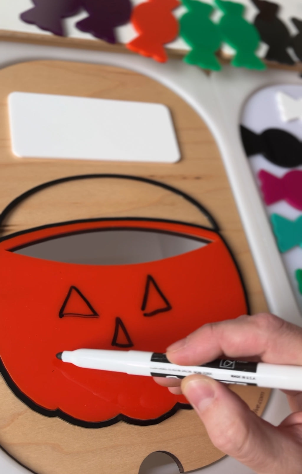 Hand drawing a jack-o'-lantern face on an orange pumpkin-shaped board with a black marker on a wooden surface