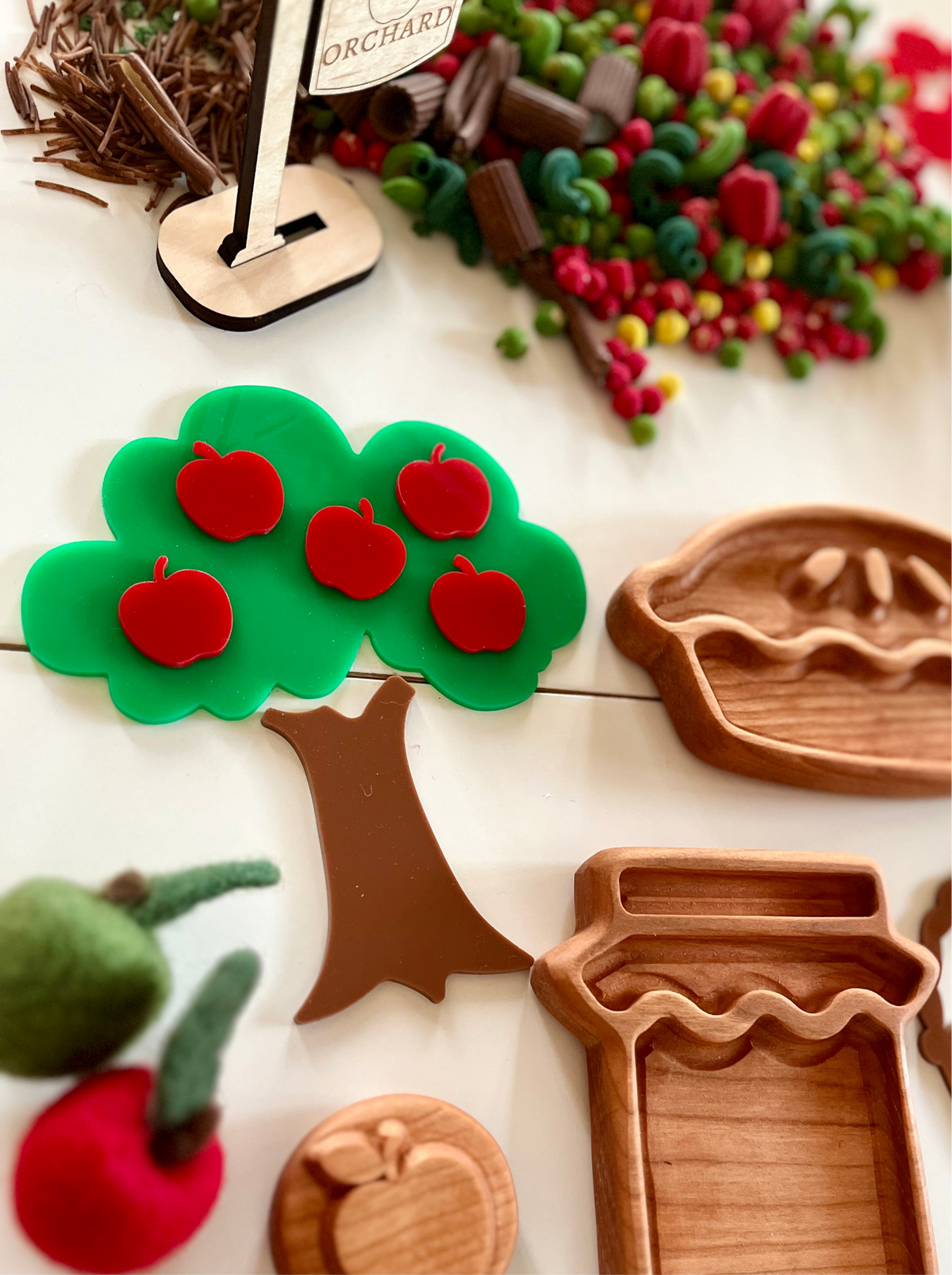 Green plastic apple tree with five red apples next to small wooden pie and jar trays on white surface
