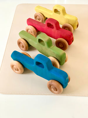 Four wooden toy trucks painted in blue, green, red, and yellow with natural wooden wheels on a beige surface