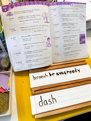 Wooden writing trays with lined white dry erase inserts displaying words from an open workbook on yellow surface