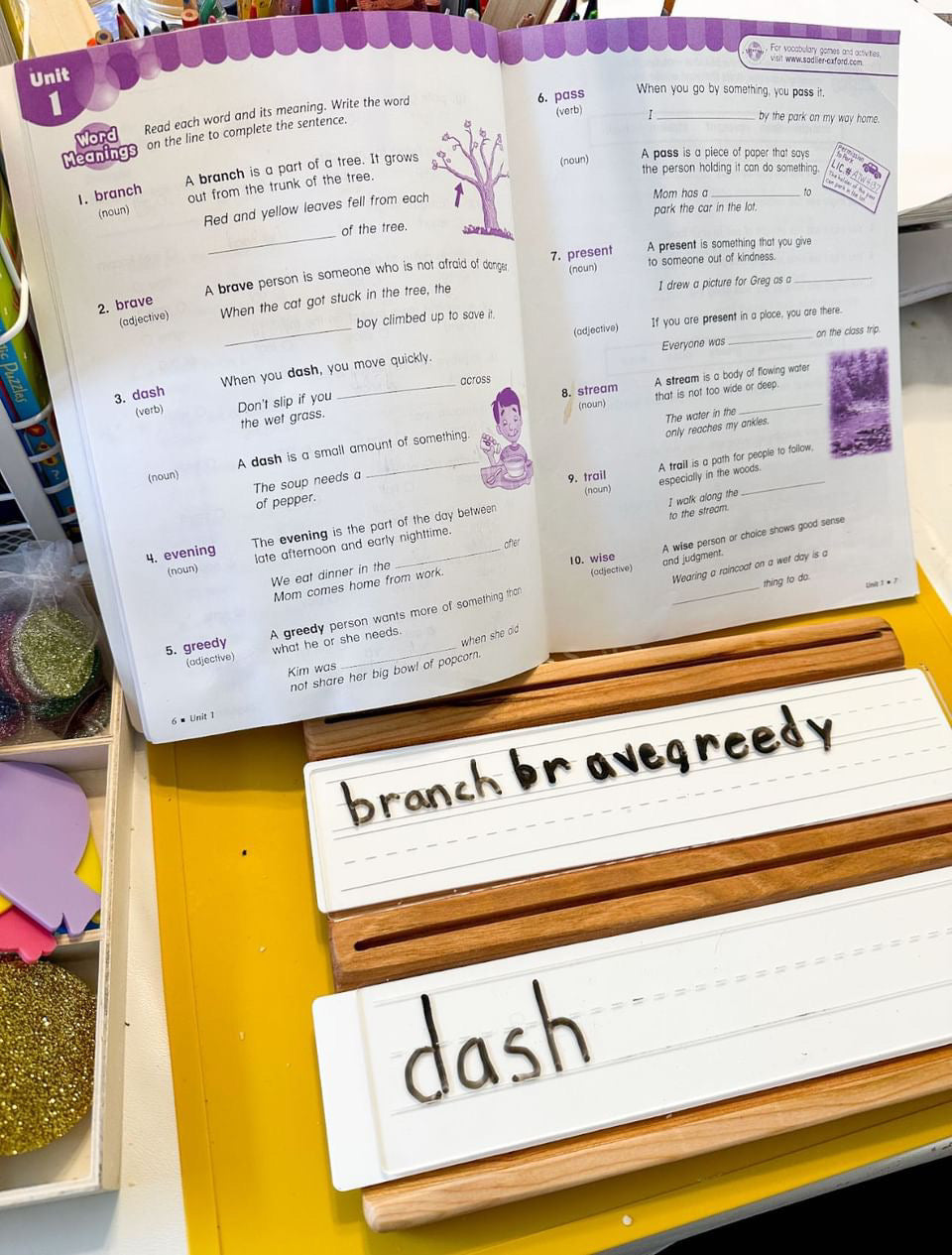 Wooden writing trays with lined white dry erase inserts displaying words from an open workbook on yellow surface