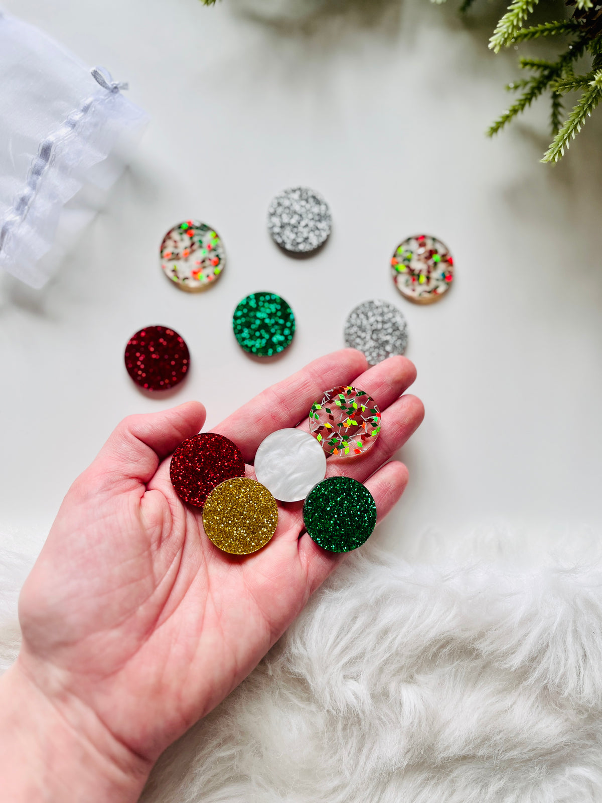 Hand holding glittery round acrylic shapes in red, green, gold, white, and confetti patterns on white surface with greenery nearby