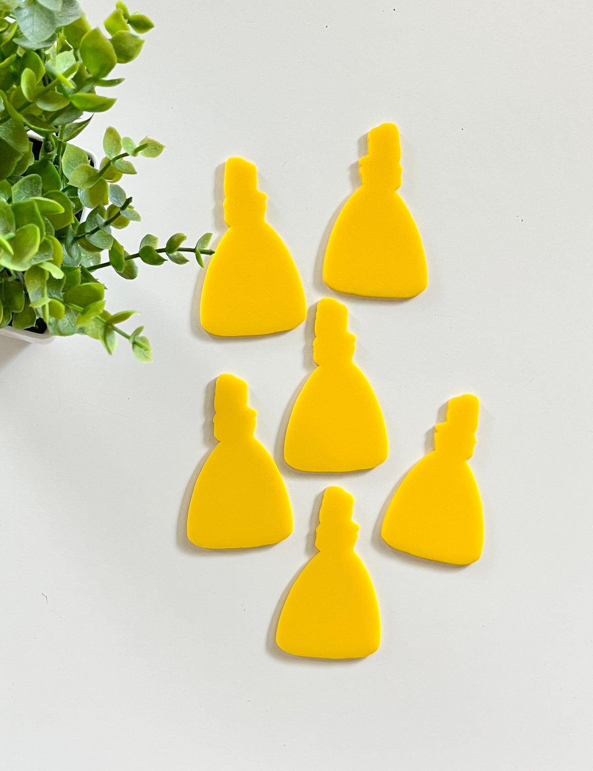 Six bright yellow dress-shaped cutouts arranged near a green leafy plant on a white surface