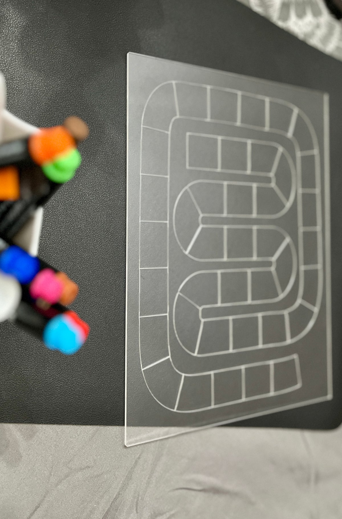 Transparent board with a white winding path grid on a black surface beside colorful marker tips in a container
