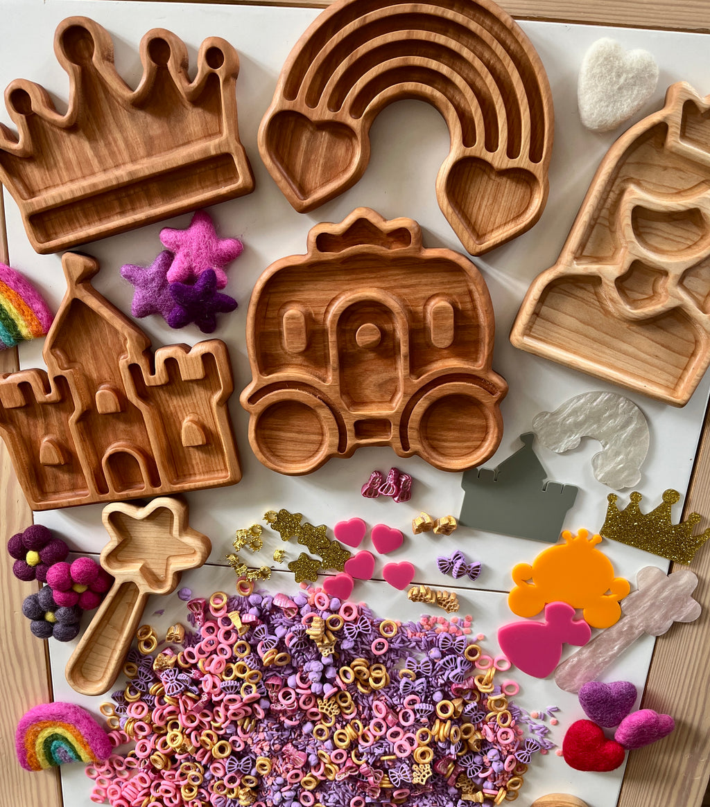 Wooden trays shaped like a crown, rainbow, castle, carriage, and house surrounded by colorful felt and small craft pieces on wood surface