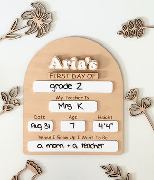 Wooden dry erase sign with customizable first day of school details surrounded by wooden floral decorations on white surface