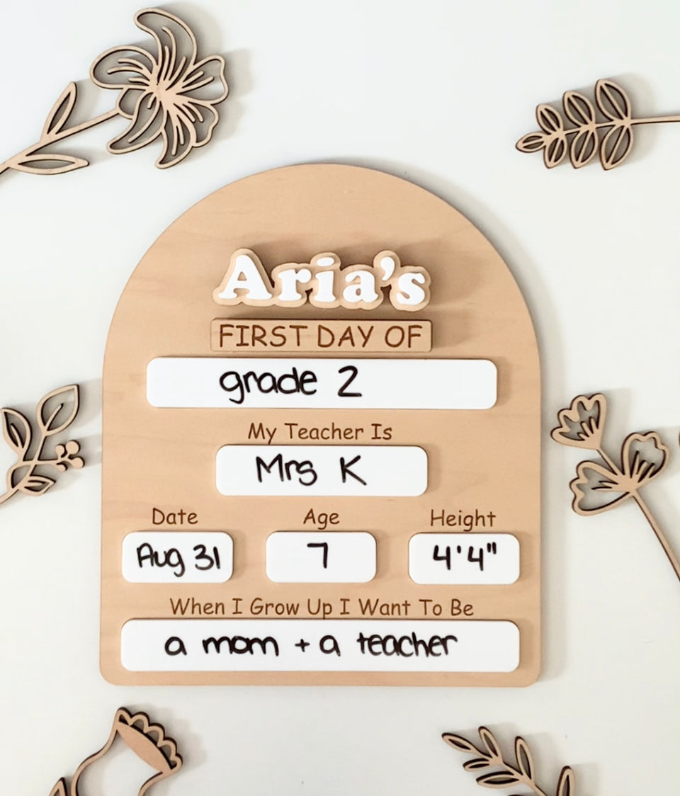 Wooden dry erase sign with customizable first day of school details surrounded by wooden floral decorations on white surface
