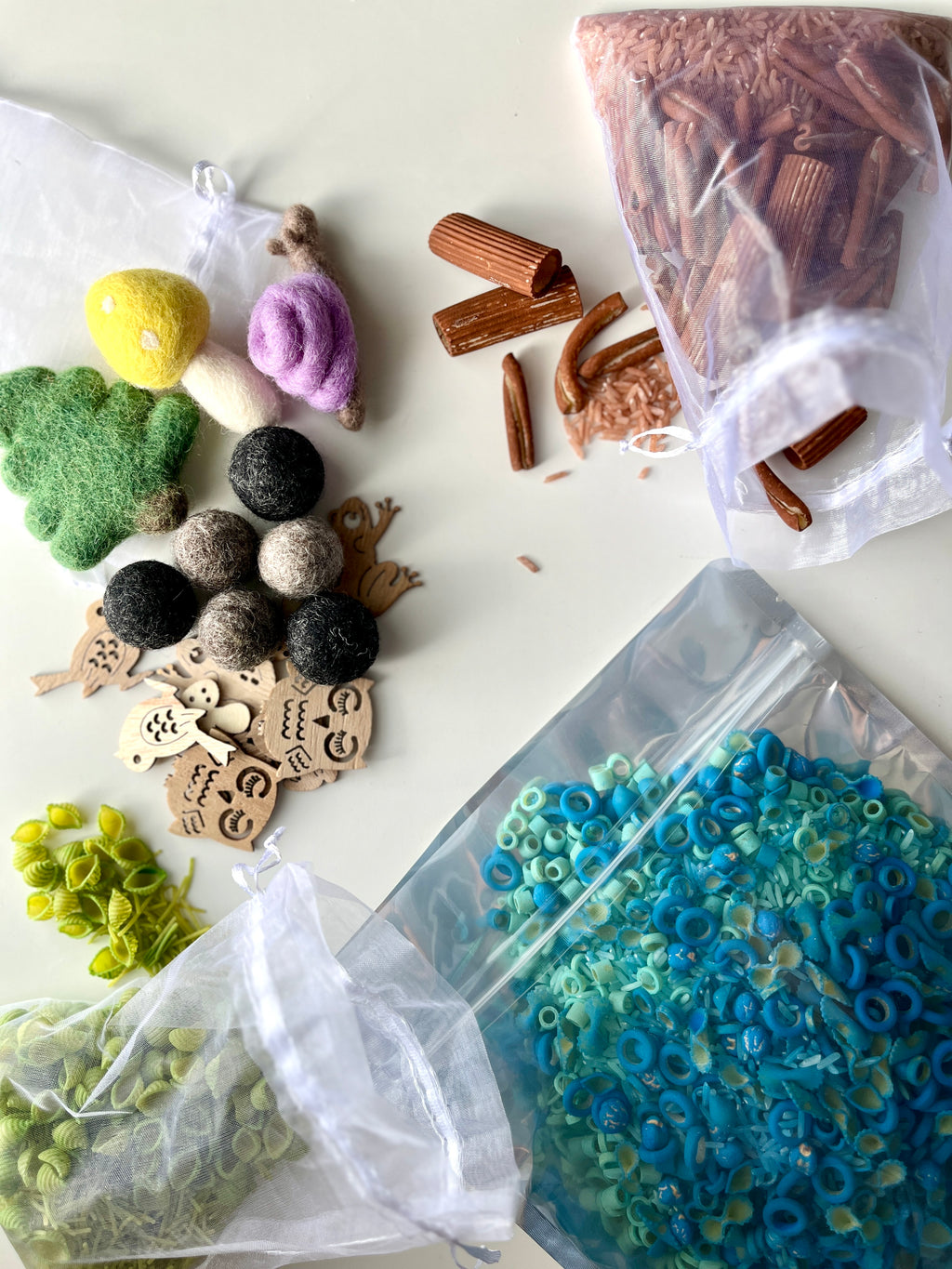 Assorted natural play materials including wooden bird shapes, felt balls, shells, green leaves, and brown pasta pieces in mesh bags