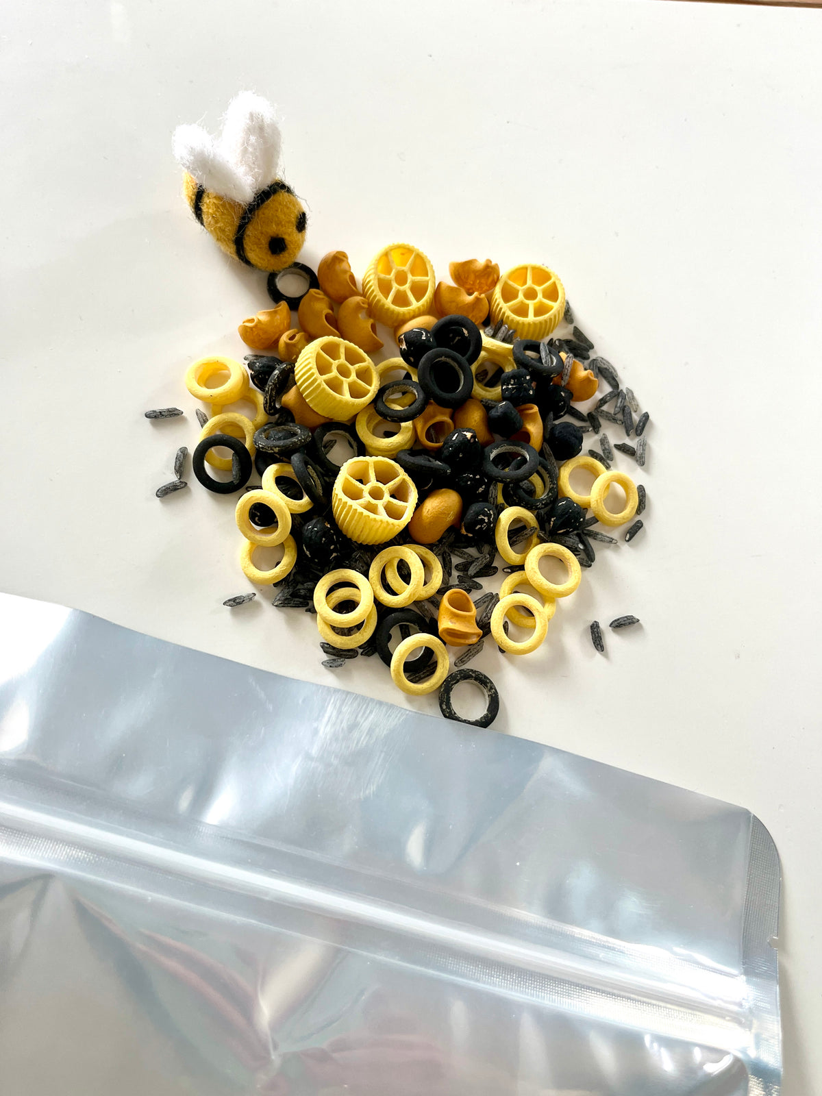 Black and yellow pasta shapes mixed with black rice grains beside a small felt bee and a partially visible silver bag