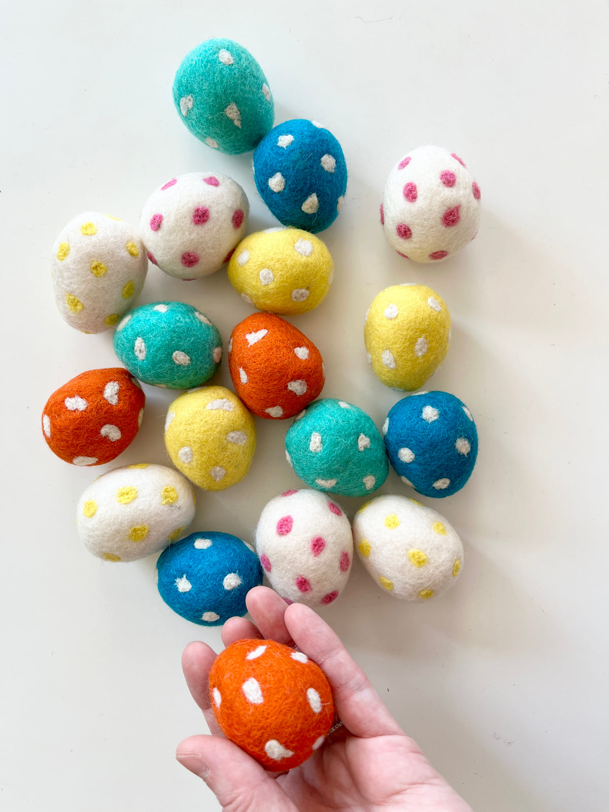 Hand holding an orange felt egg with white dots among scattered felt eggs in blue, yellow, white, and teal with polka dots on white surface