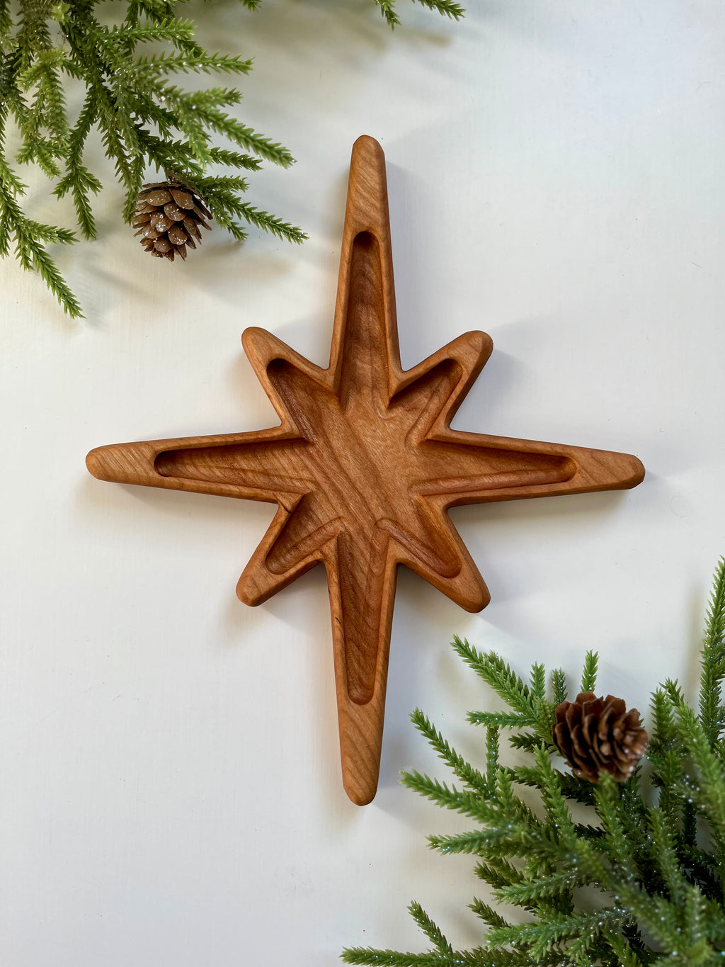 Carved wooden star tray with multiple points surrounded by pine branches and pine cones on white surface