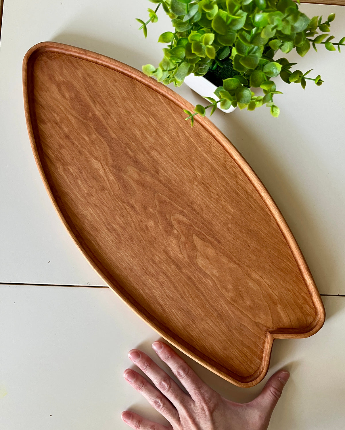 Handcrafted wooden tray shaped like a fish with visible wood grain next to a small green potted plant on white surface