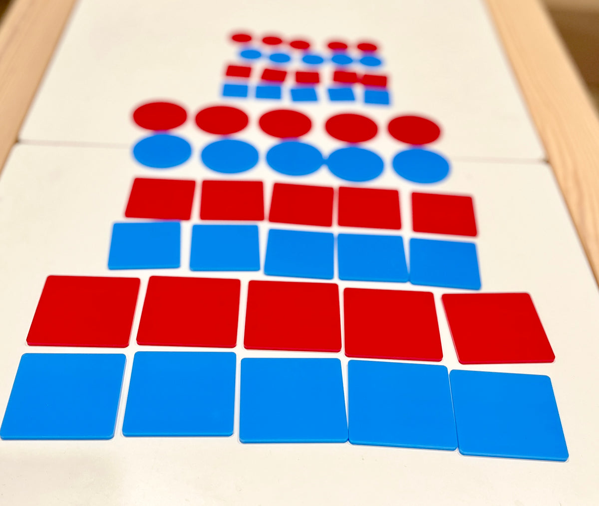 Red and blue flat circular and square plastic tiles stacked in alternating rows on a white surface with a wooden frame