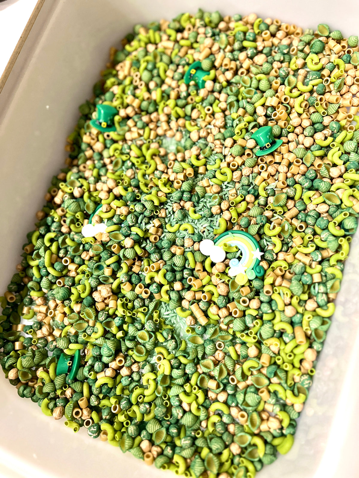 Container filled with green and beige pasta shapes, small beads, and a rainbow and green hat charms scattered inside