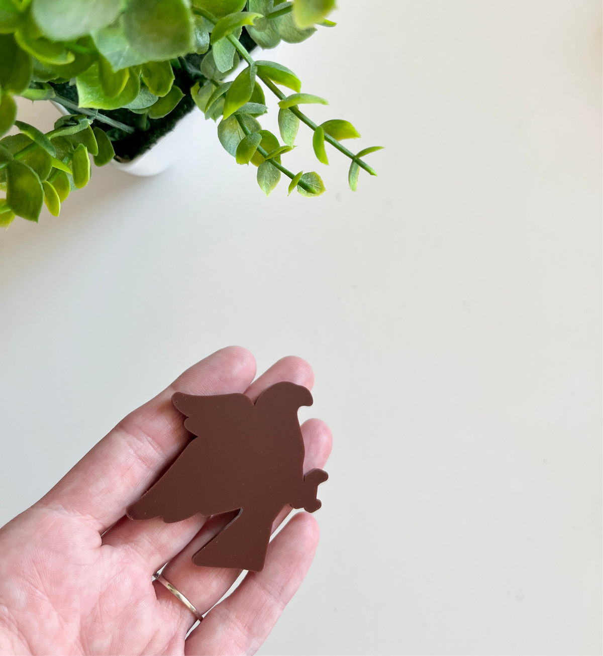 Hand holding a smooth dark brown bird-shaped cutout with a green leafy plant in a white pot above on a light surface