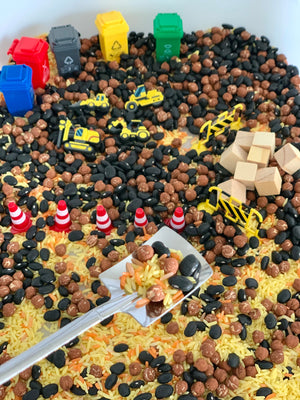 Miniature construction vehicles, traffic cones, and wooden blocks placed on a mix of beans, rice, and pasta in a sensory bin