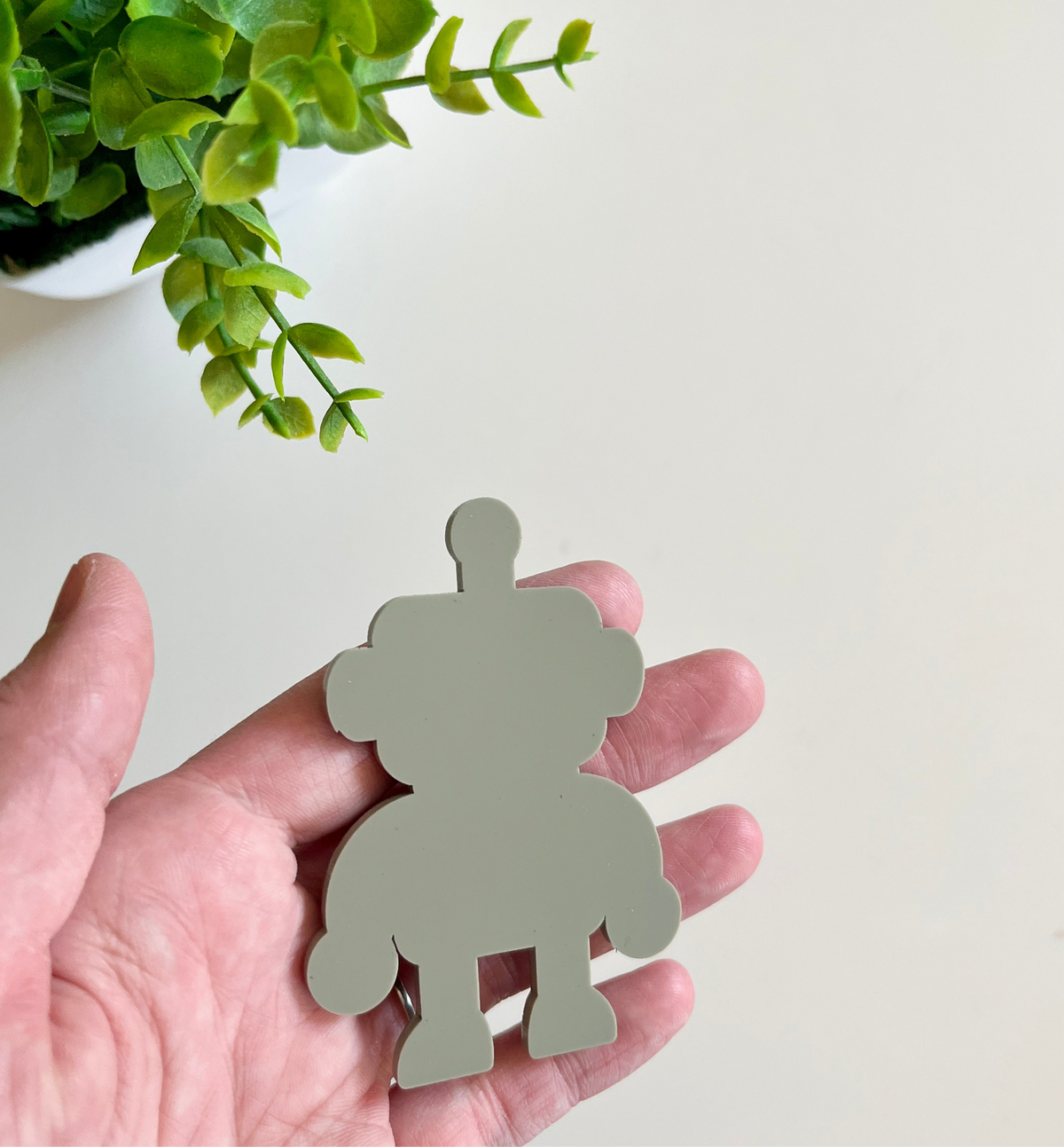 Hand holding a smooth light gray robot-shaped cutout with a green leafy plant in a white pot nearby