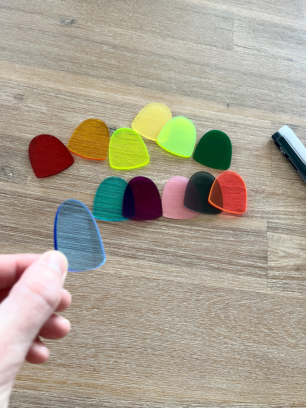 Hand holding a blue translucent gum drop piece with nine additional colorful translucent gum drops arranged on a wood surface near a black and white dry erase marker, showcasing Little Dry Erasables — Set of 10 Translucent Gum Drops, handcrafted homeschool education resources.