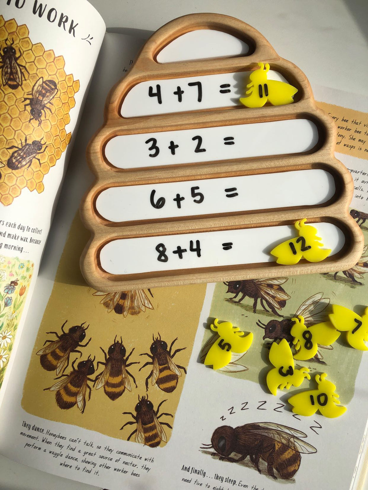 Wooden beehive-shaped board with math addition problems and yellow bee-shaped number tokens on illustrated bee book pages