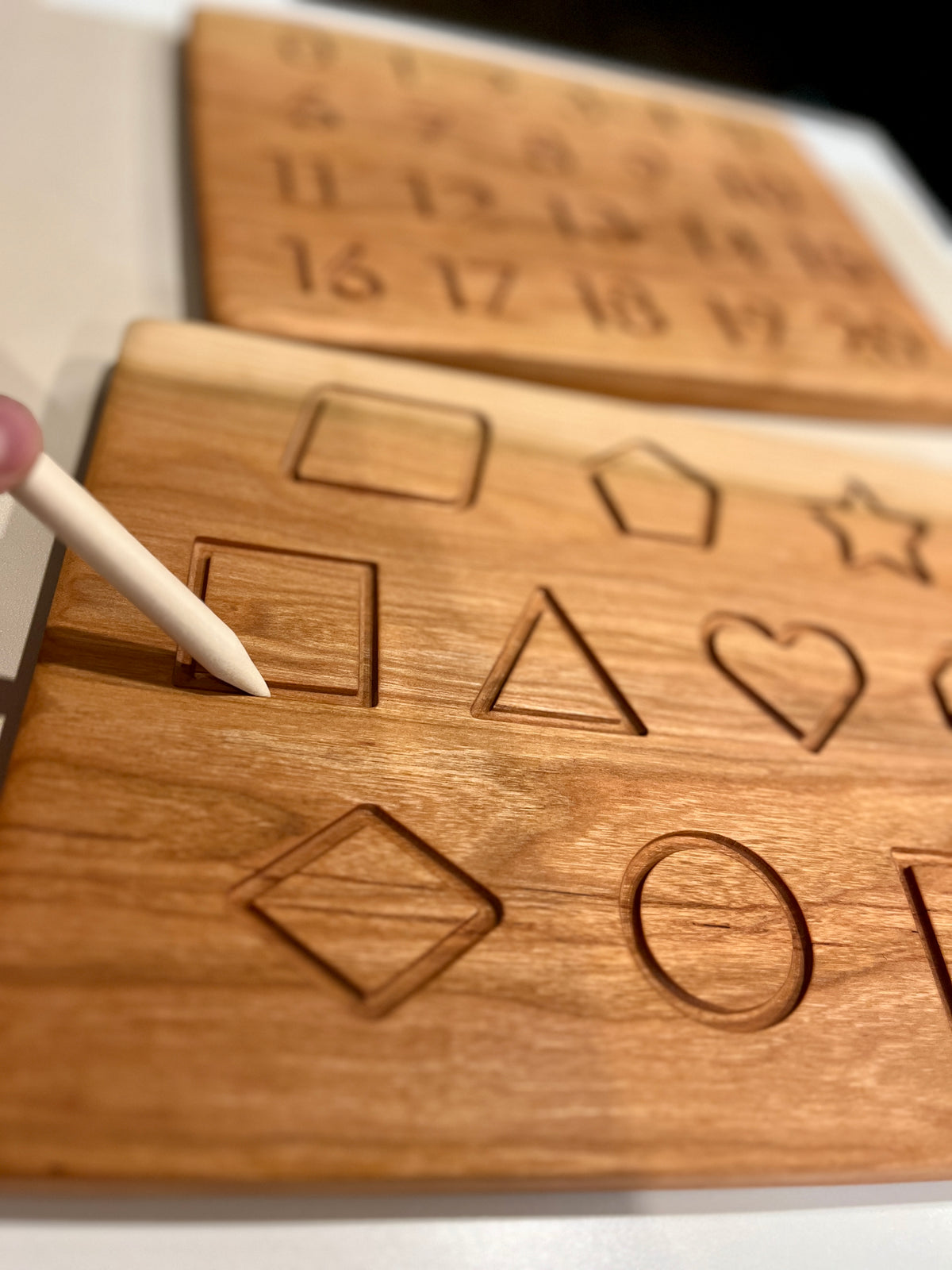 Wooden shape tracing board with engraved geometric shapes and a wooden stylus pointing at a square indentation