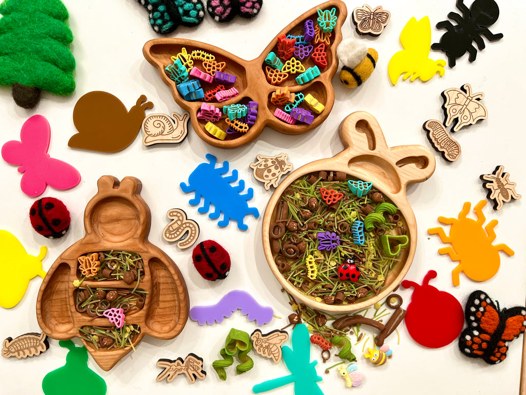 Wooden bug-shaped trays filled with colorful small plastic bugs and natural dried grass surrounded by felt and wooden insect shapes