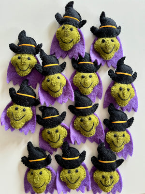 Multiple small handmade felt witch faces with green skin, black hats, and purple collars arranged on a light surface.