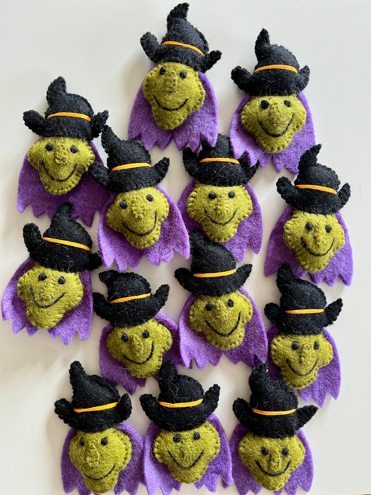 Multiple small handmade felt witch faces with green skin, black hats, and purple collars arranged on a light surface.