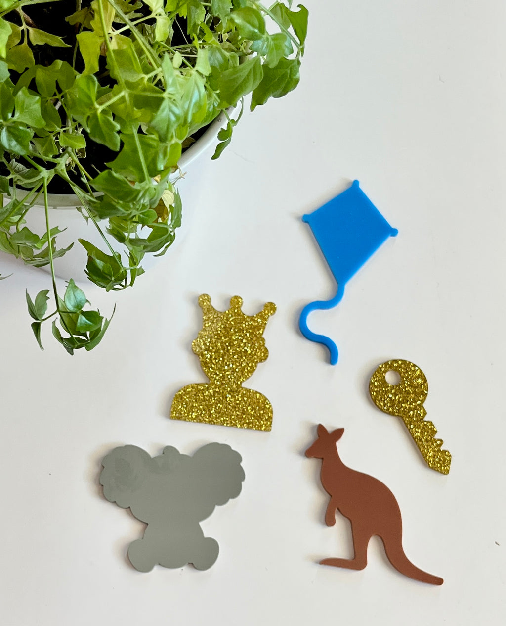 Five colorful acrylic shapes including green plant, gold crown silhouette, blue kite, gold key, gray koala, and brown kangaroo on white surface