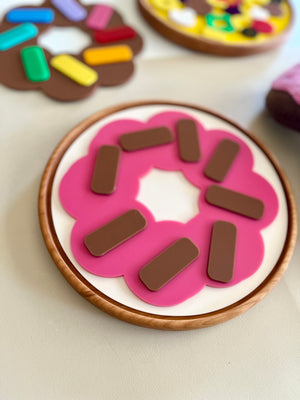 Wooden toy donut with pink icing and rectangular brown sprinkles on a light surface with other toy pastries nearby