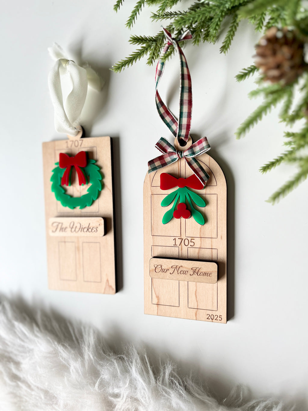 Two wooden door-shaped holiday ornaments decorated with bows, greenery, and personalized plaques hanging on fabric ribbons