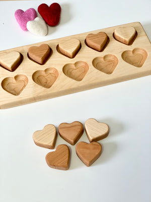 Wooden heart-shaped blocks in two tones with matching recessed slots on a light wood tray, plus three felt hearts nearby.