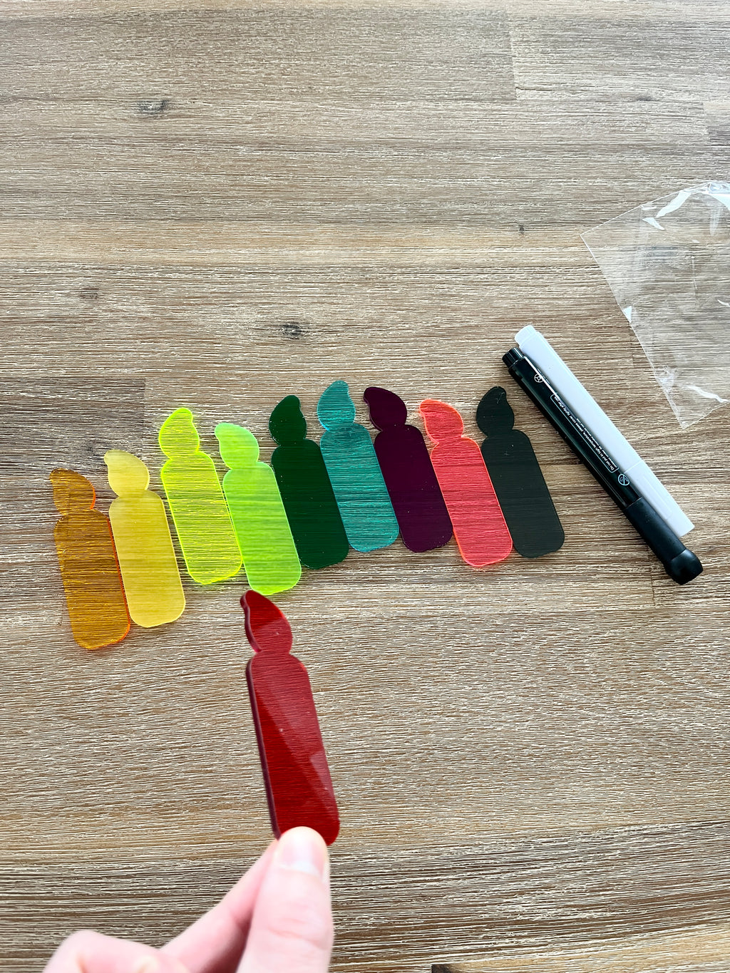 Hand holding a red translucent candle piece with nine additional colorful translucent candles and black and white dry erase markers on a wood surface, showcasing Little Dry Erasables - Translucent Candles - Set of 5 or 10, handcrafted homeschool education resources.