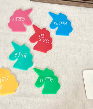 Brightly colored unicorn-shaped acrylic pieces with handwritten math problems on a fabric surface, part of Little Dry Erasables - Set of 7 Large Unicorns, handcrafted homeschool education resources.
