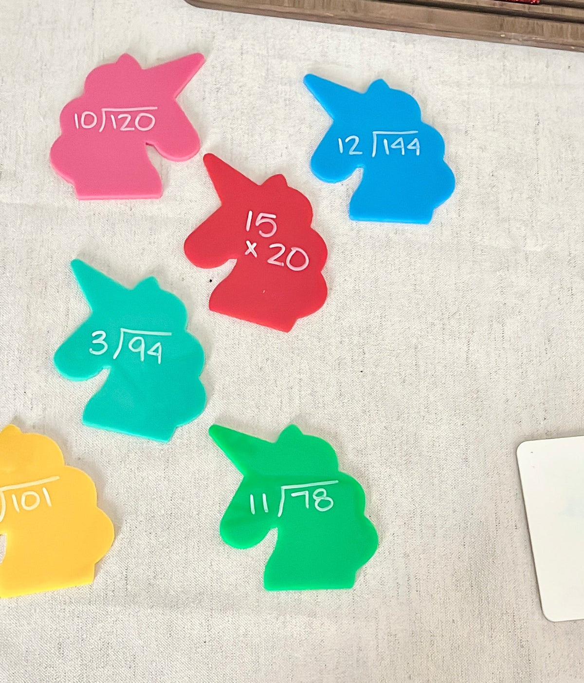 Brightly colored unicorn-shaped acrylic pieces with handwritten math problems on a fabric surface, part of Little Dry Erasables - Set of 7 Large Unicorns, handcrafted homeschool education resources.