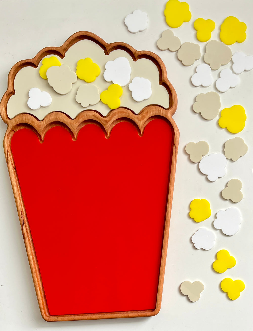 Wooden tray with scalloped edge and red base filled with yellow, white, and beige popcorn-shaped tokens on white surface