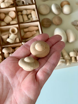 Hand holding two round wooden knob shapes with assorted unfinished wooden pieces in a compartmented box in the background