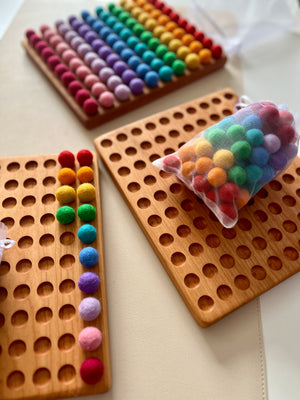 Bag of colorful felt balls next to wooden hundred boards with felt balls arranged by rainbow colors