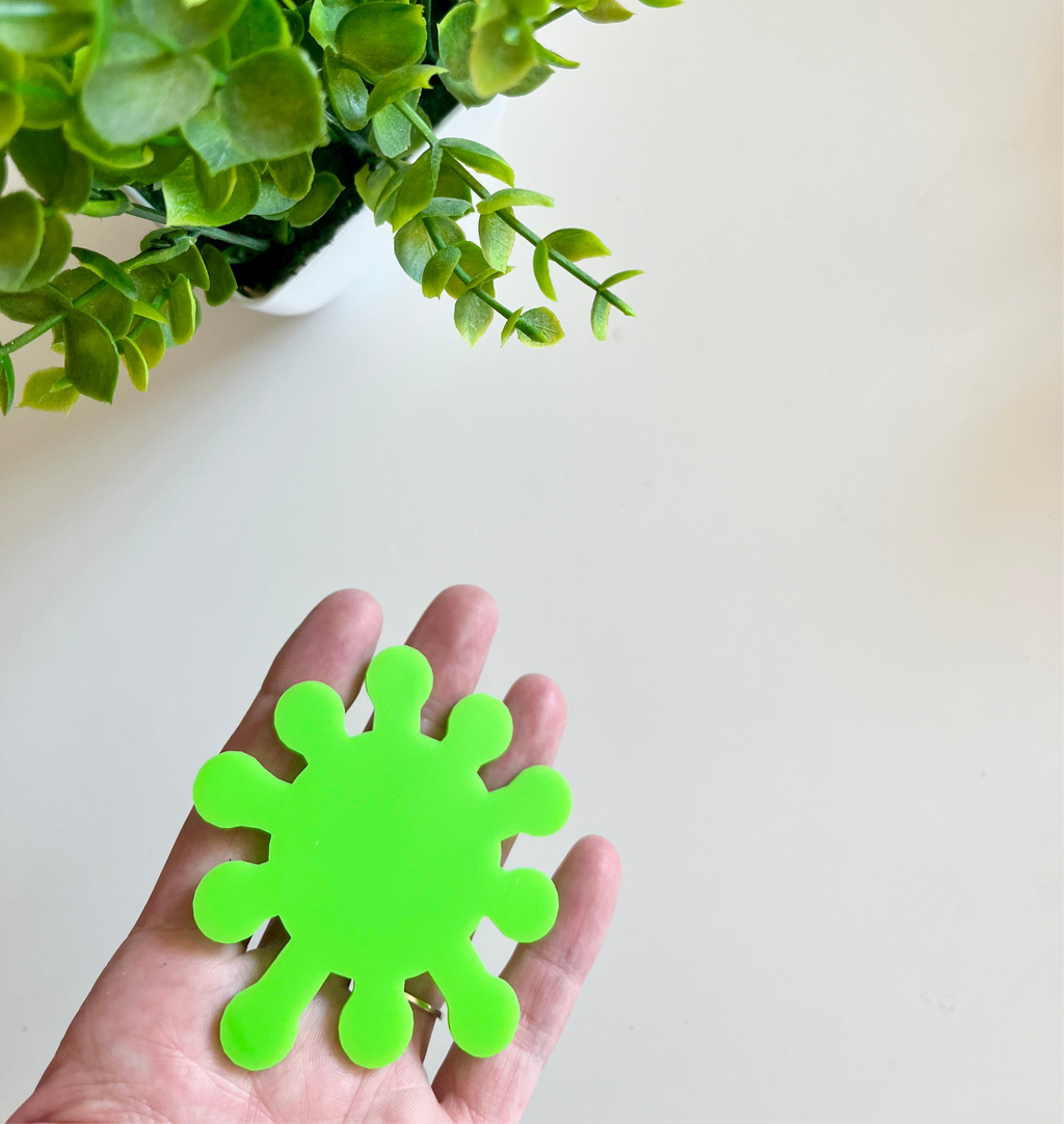 Hand holding a bright green smooth cutout shaped like a virus with green leafy plant above on a white surface